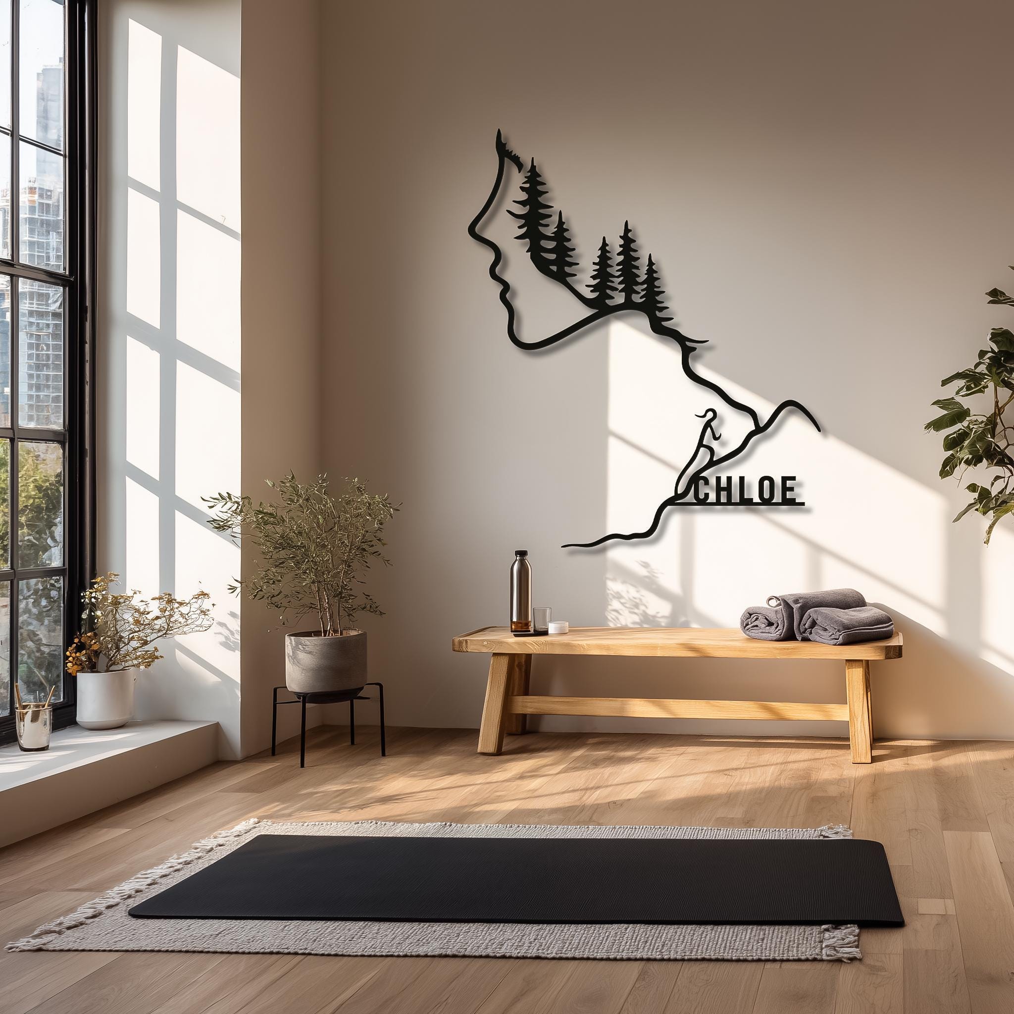 Personalized black metal mountain climber wall art with woman’s profile, pine trees and custom name “CHLOE” on the trail, styled in a yoga studio with bench and mat. Custom hiking metal wall art, name sign, forest line art decor, unique adventure lover gift idea.
